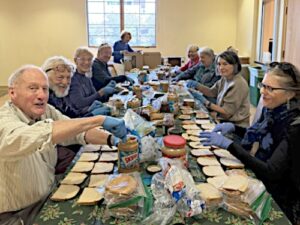 About a dozen people sitting at table making sandwiches