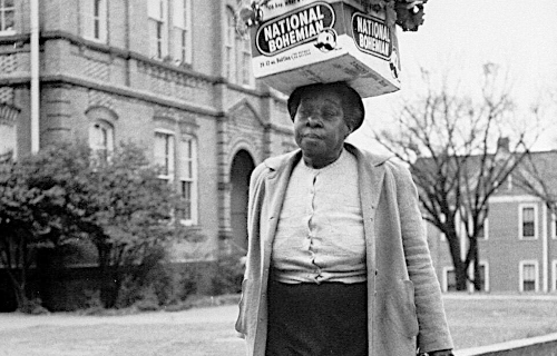 Woman walking on sidewalk carrying box of turnips on her head