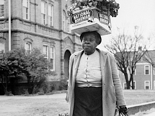 Woman walking on sidewalk carrying box of turnips on her head